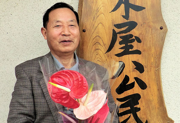 公民館の看板の前で赤やピンクの色をした観葉植物を手に笑顔を見せる廣野さん