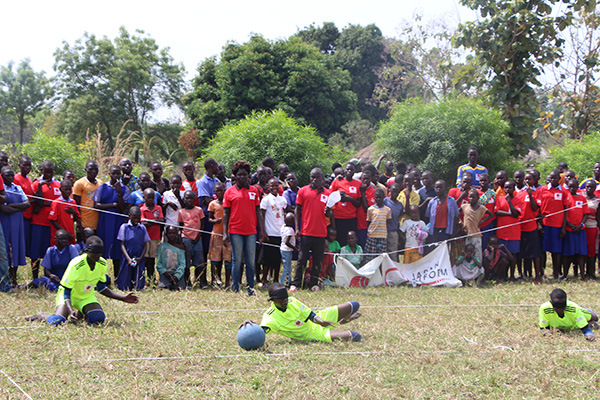 3人の選手が地面の上で間隔をあけながら、ボールを横になって持っていたりかまえている。背後には観客が立って応援している
