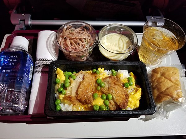 機内食のプレートには、ペットボトルに入った水、プラスチックカップに入ったお蕎麦、お茶、パンが置かれている。ご飯の上には、鶏肉、卵、グリーンピースが載せられている