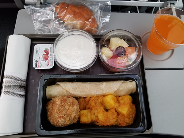 機内食のプレートには、パン、ジャム、パイナップルなどが入ったカットフルーツ、ジャガイモや揚げ物、ロール状に巻かれたチャパティ、ジュースなどが並んでいる