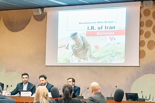 壇上に3名のスピーカーが並びスクリーンにイランのプレゼンスライドの表紙が映されている。地雷除去員の写真