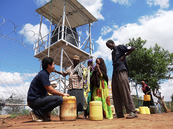 給水塔の下で、水のポリタンクを手に笑顔を見せる人々