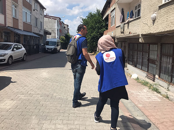 AARスタッフの男性と女性が、難民世帯を訪れるため、民家前の路上を歩いている。