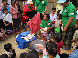 沐浴の方法を伝えている。大きなたらいの横で赤ちゃんを抱いている女性のまわりに、村人が集まる