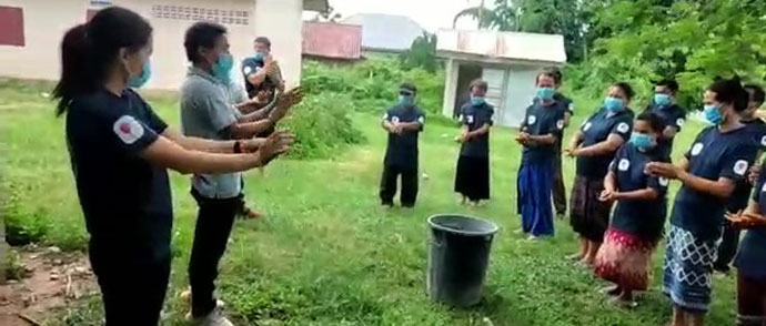 屋外で前に立った2人が手洗い方法を伝えている。それを10名ほどの参加者が真似て手洗いの練習をしている。全員マスクをしている。