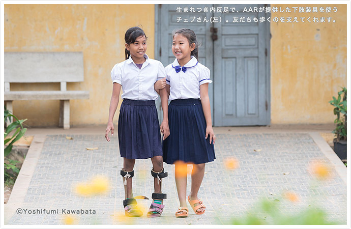校舎内を、女の子2人が笑顔で歩いている　一人の子は足が不自由で、両足に補助具をつけており、もう一人の子が、その子の歩行を支えるように腕を組み、歩いている
